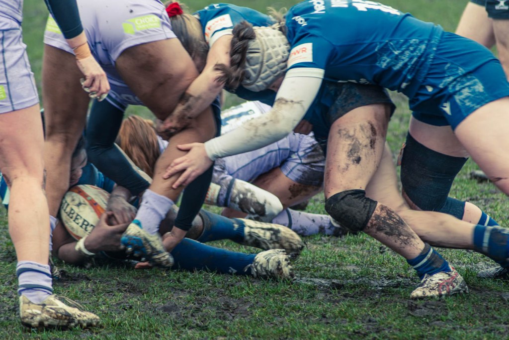 A tense moment in a women's rugby match, with players in muddy uniforms competing for the ball on the grass.