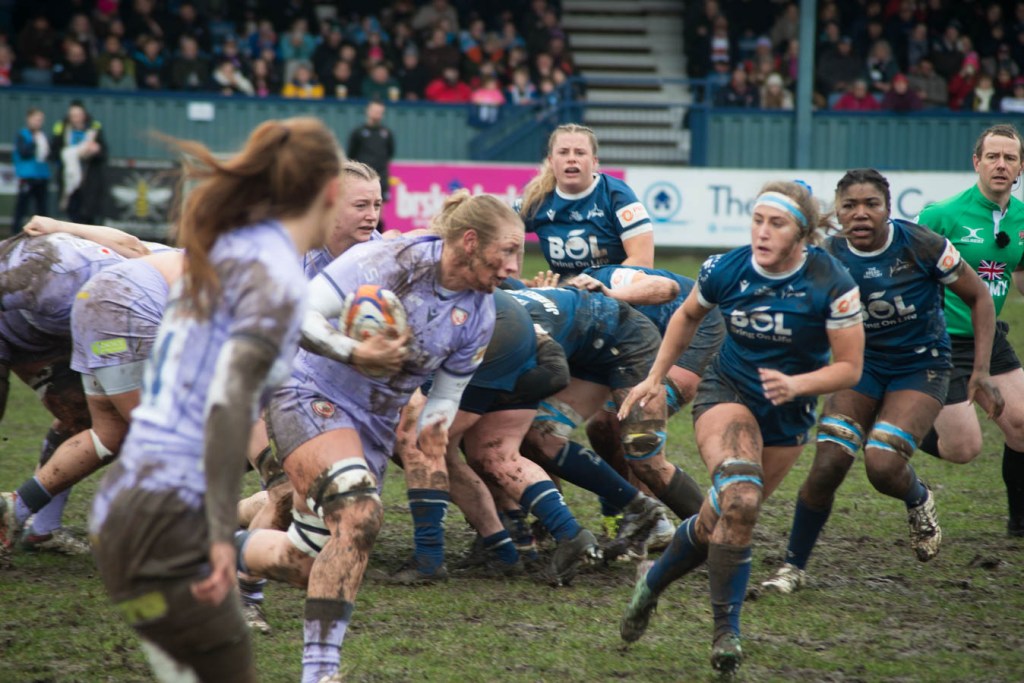 A rugby match in muddy conditions featuring female players in blue and purple uniforms, intensely competing for the ball during a tackle.