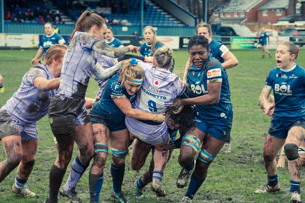Female rugby players in a muddy field engaged in a challenging tackle during a match, showcasing physical competitiveness and teamwork.