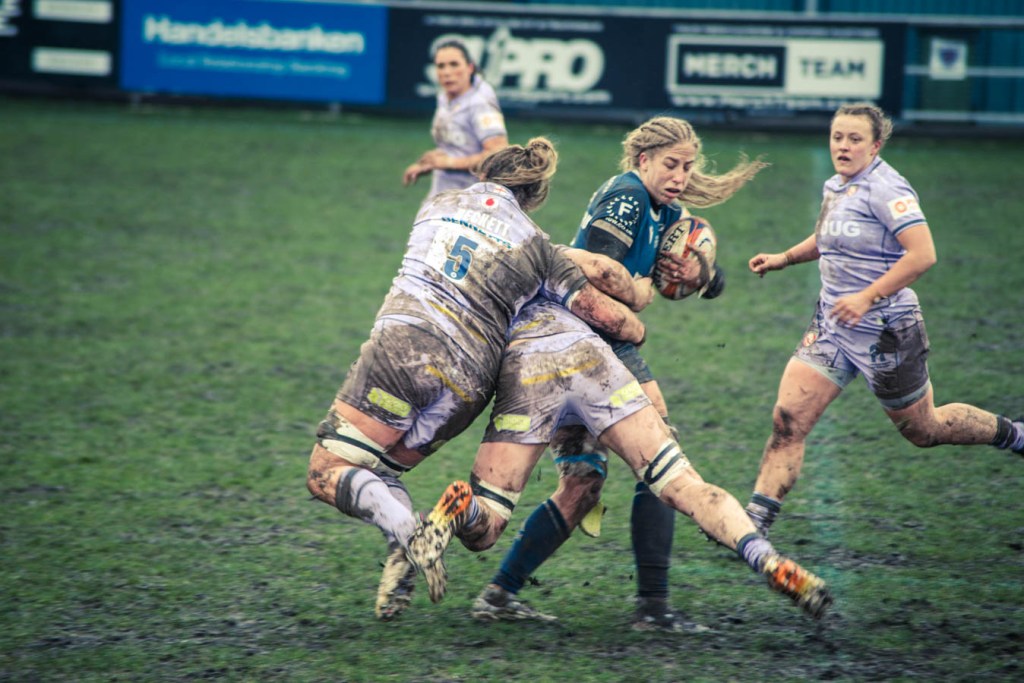 A female rugby player in a blue jersey is evading two opponents while carrying the ball, with muddy uniforms and a grassy field in the background.