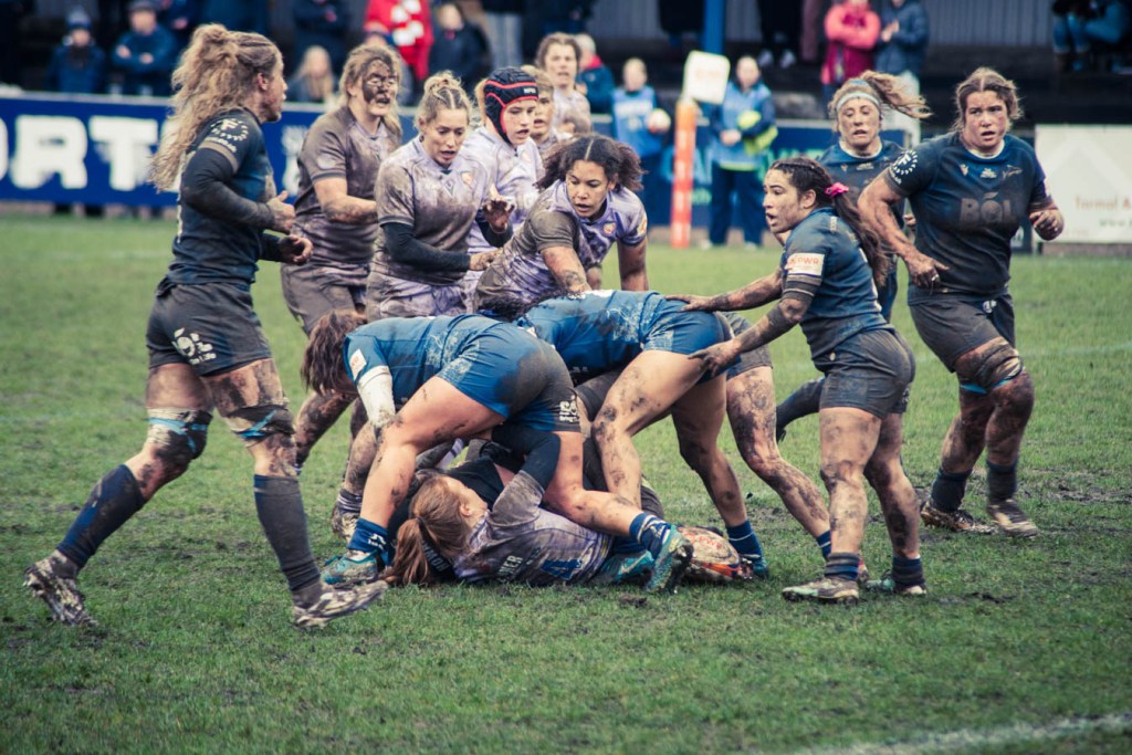 A women's rugby match in progress, with players in muddy uniforms engaged in a tackle on a grassy field.