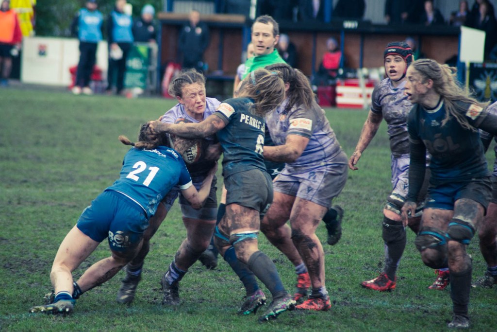A rugby match in muddy conditions featuring players in action, with one player in blue number 21 tackling an opponent from the opposing team.
