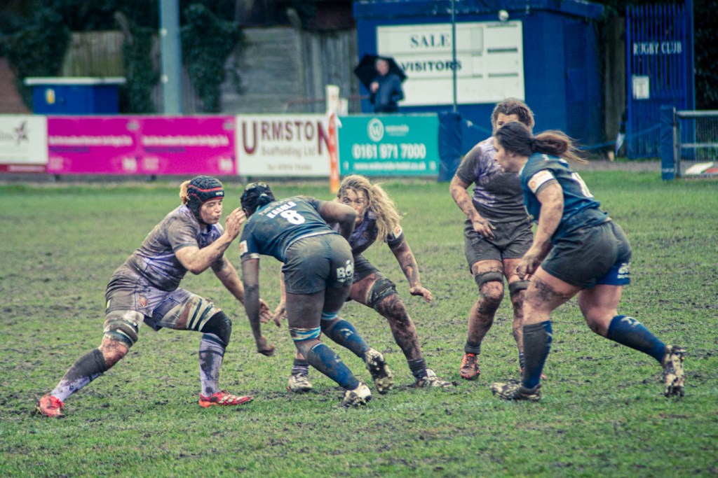 A rugby match in progress with players in muddy uniforms engaged in a tackle on a grassy field.