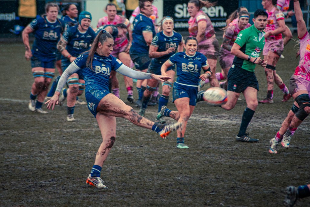 A female rugby player in blue uniform is kicking a rugby ball while surrounded by teammates and opponents during a muddy match.