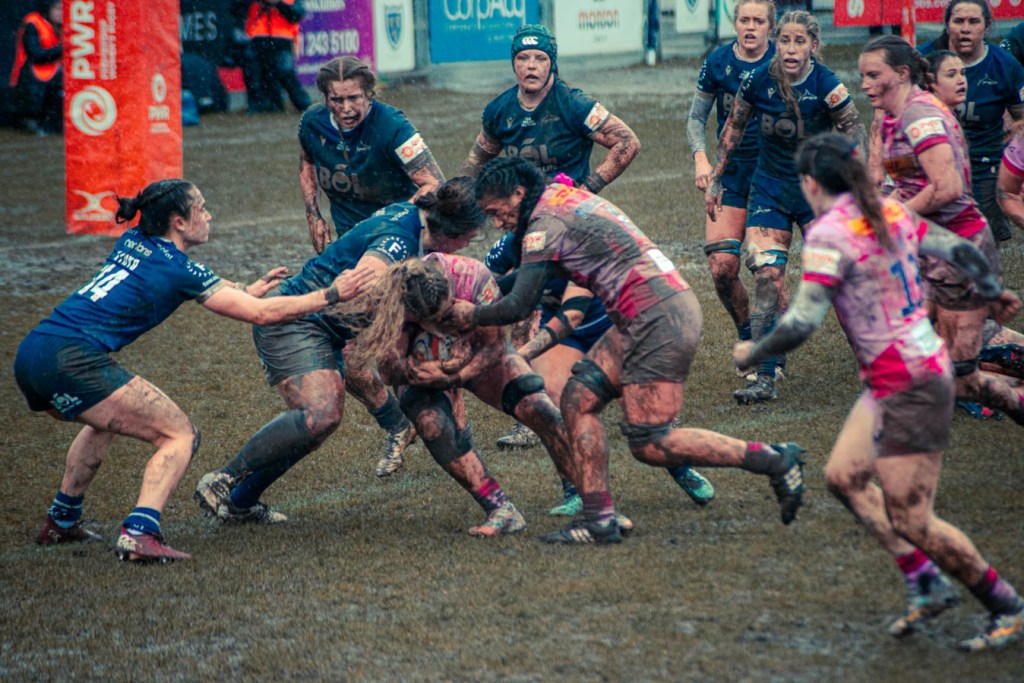A rugby match in progress on a muddy field, featuring several female players in action, tackling a player with the ball amid rain and challenging conditions.