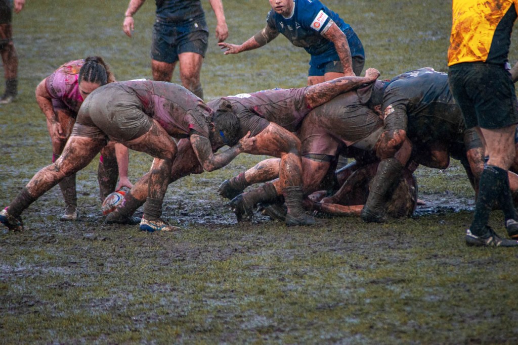 A rugby match in muddy conditions, featuring players from both teams engaged in a scrum, with mud splattered on their uniforms.