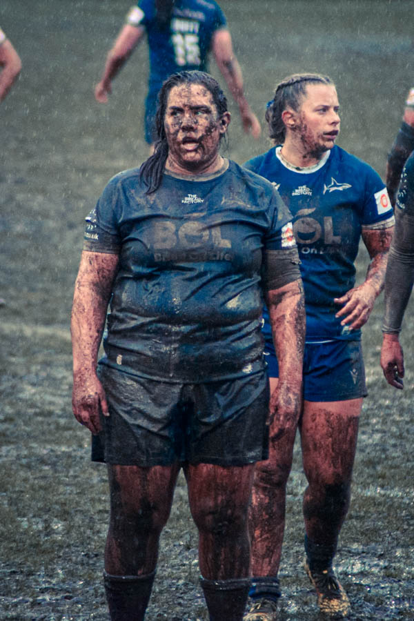 Two rugby players in muddy uniforms during a match, with one player prominently covered in mud while standing in the foreground.