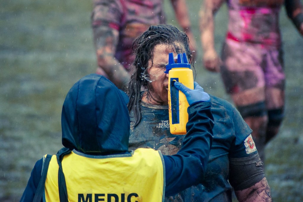 A medic in a yellow vest sprays water into the face of a muddy athlete during a rainy outdoor event, with other participants visible in the background.