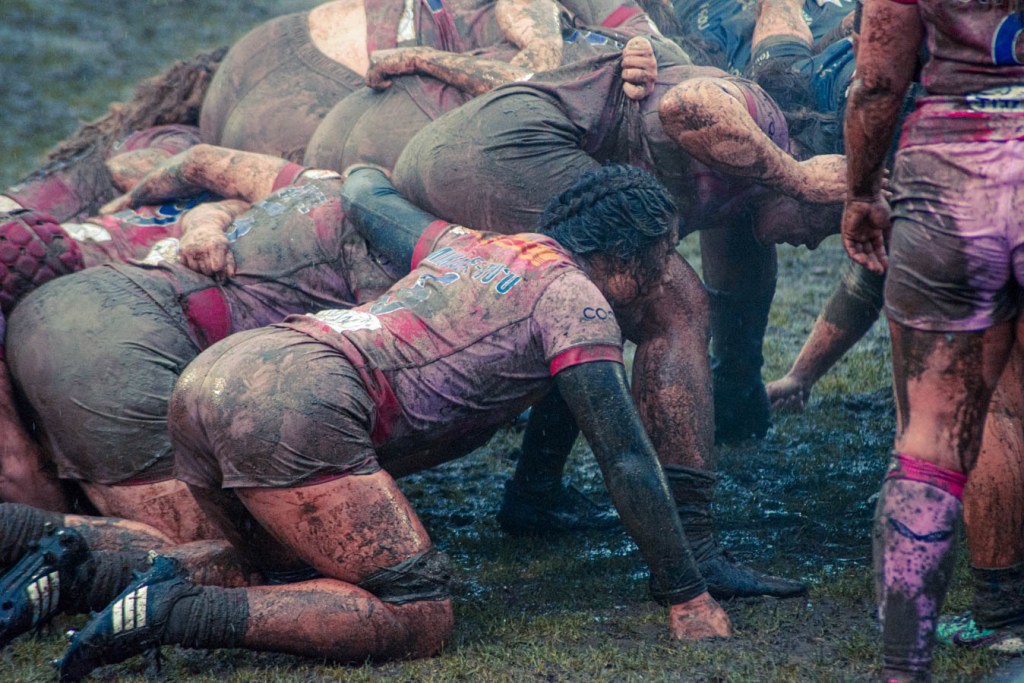 A close-up view of a rugby scrum, featuring players in muddy uniforms exerting strength and teamwork in a wet and muddy field.
