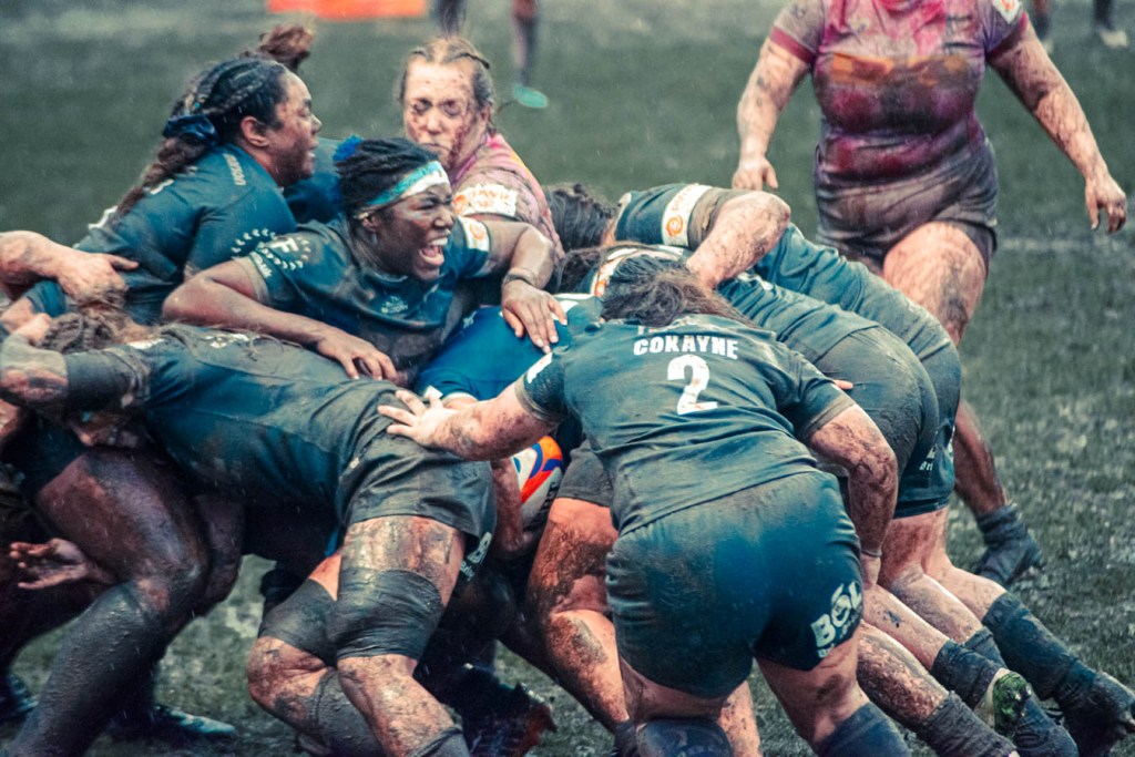 A group of female rugby players engaged in a maul, covered in mud on a grass field, showing intense teamwork and determination.