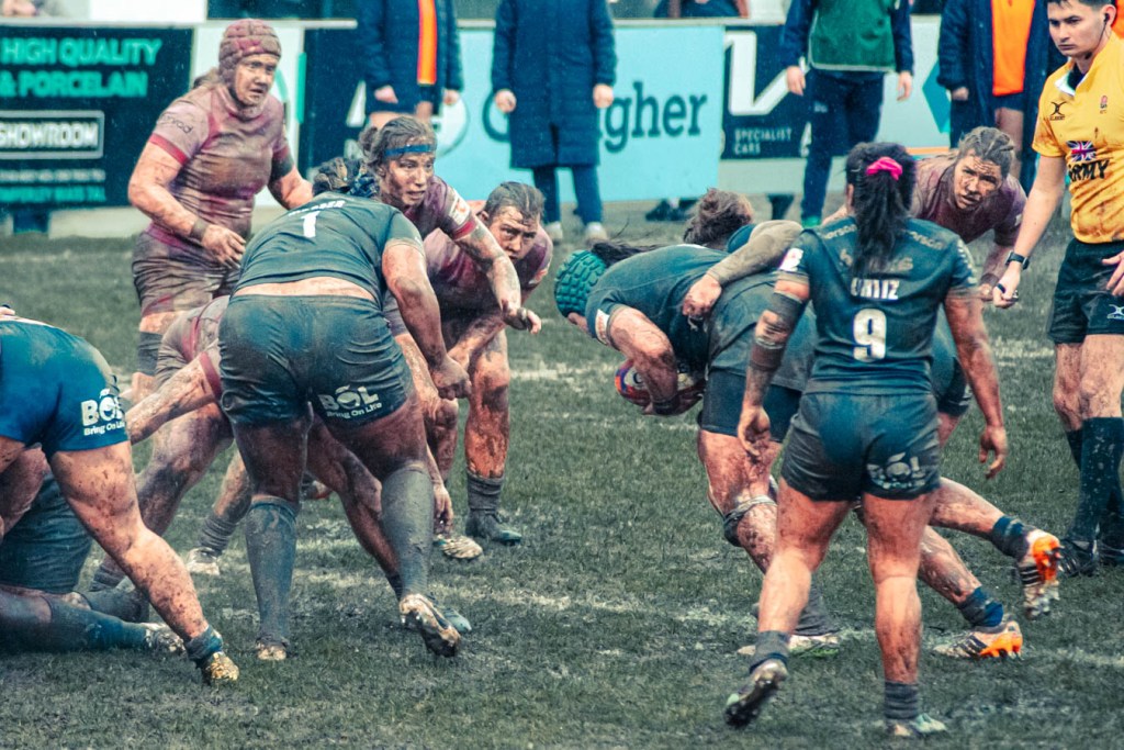 A rugby match in muddy conditions, featuring players in dark jerseys engaged in a tackle. Two players are in focus, one preparing to pass the ball while others surround them. The field is muddy, and the spectators are blurred in the background.