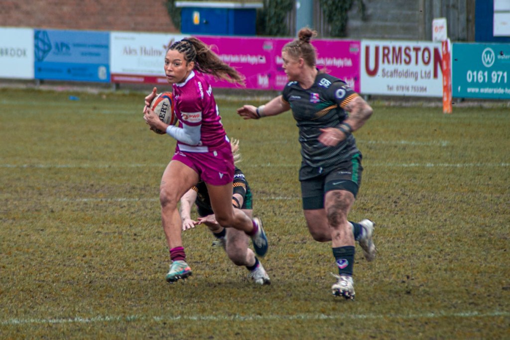 A rugby player in a pink uniform is sprinting across a muddy field while holding a rugby ball, with an opposing player in a dark uniform attempting to tackle her from behind.