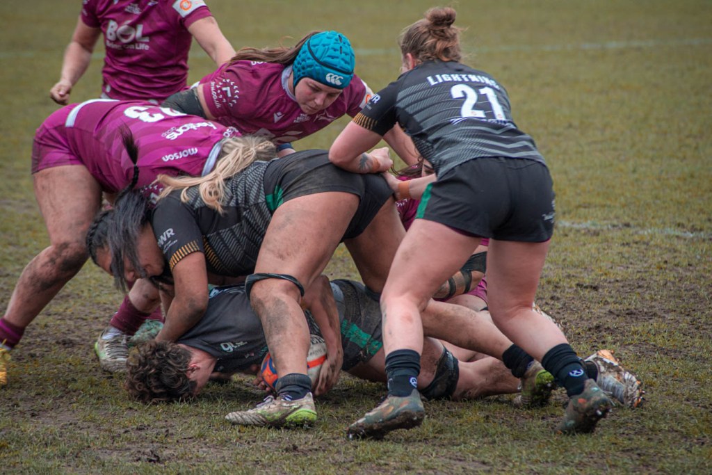 A women's rugby match in muddy conditions, featuring players in maroon and black uniforms engaging in a tackle on the field.