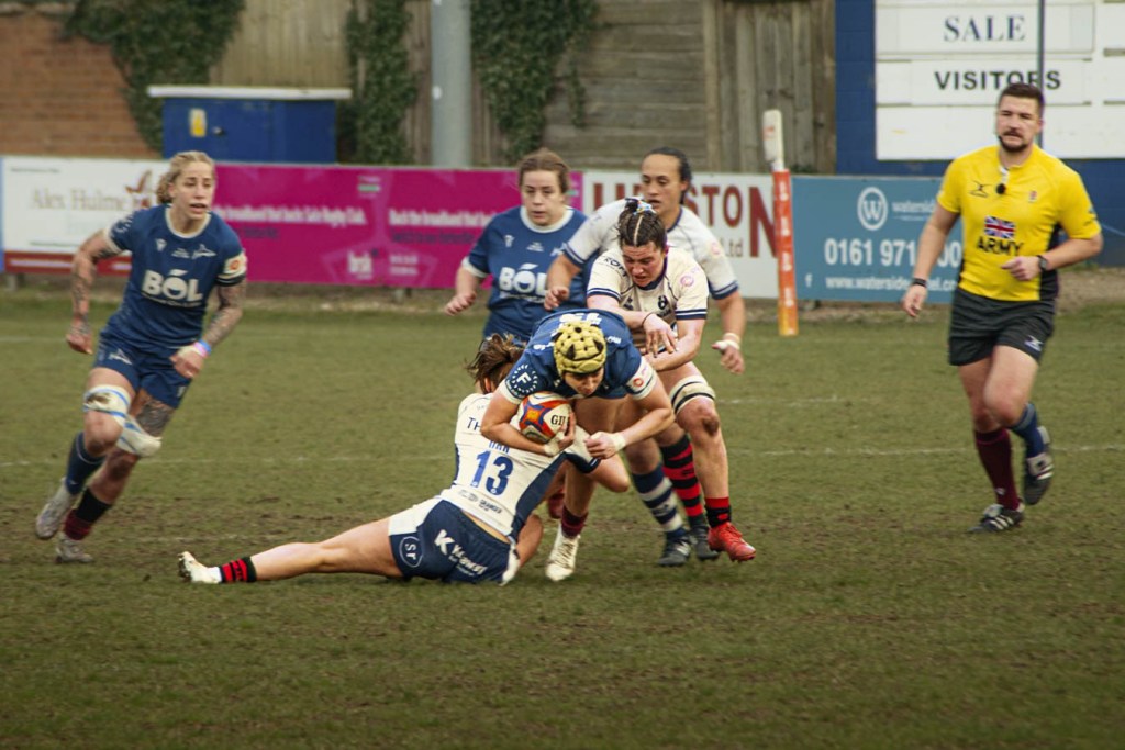 A women's rugby match in action, with players from opposing teams engaged in a tackle on a muddy field.