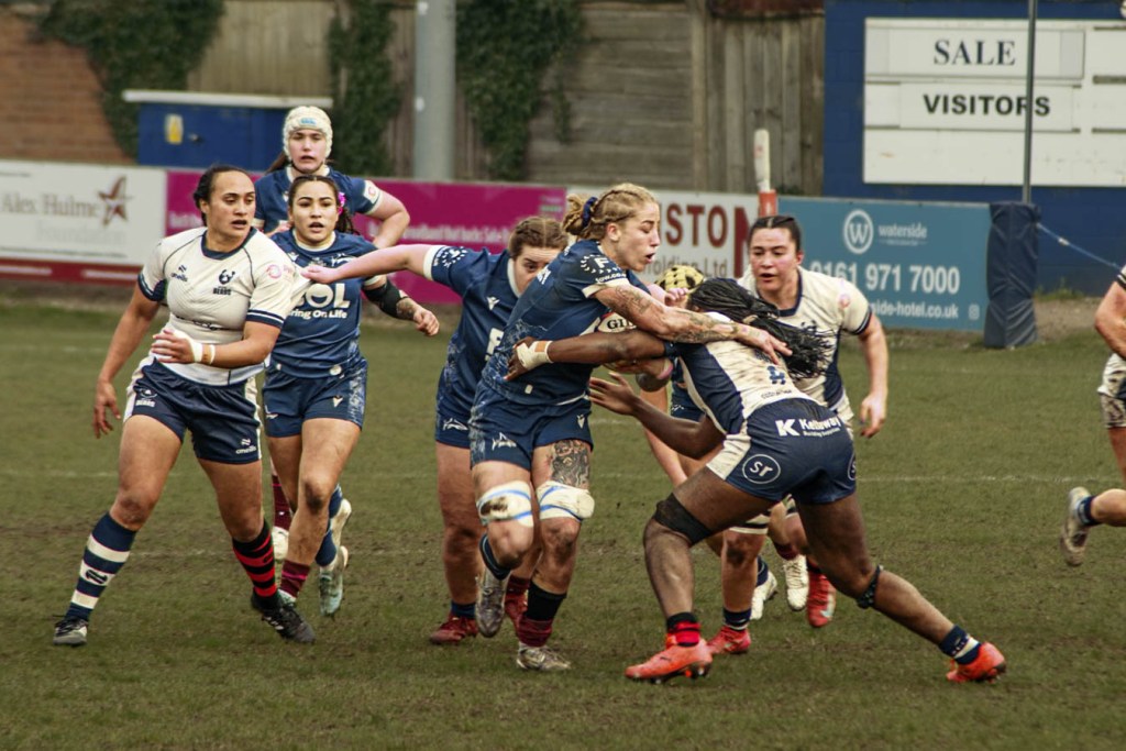 A rugby match in action, with players from two teams engaged in a tackle. The players are wearing different colored uniforms, one team in dark blue and the other in white. The scene captures the intensity of the game on a grassy field.