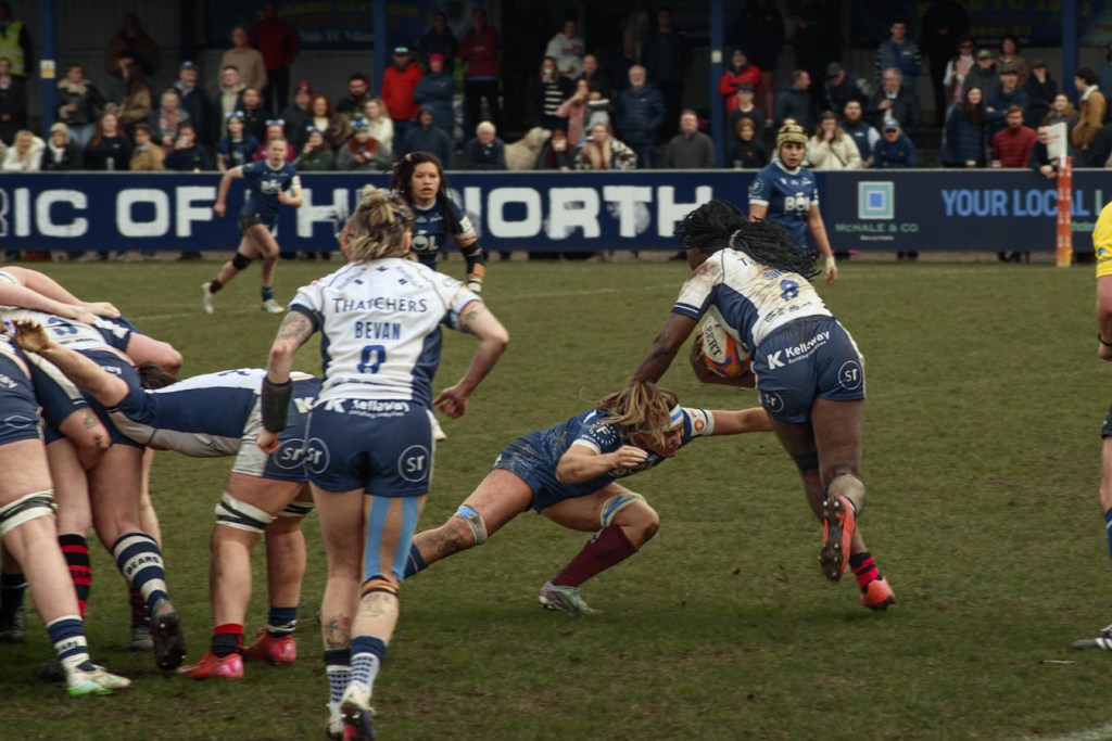 A women's rugby match in action, with a player in blue attempting to evade a tackle while carrying a rugby ball, surrounded by teammates and opponents on a grassy field.