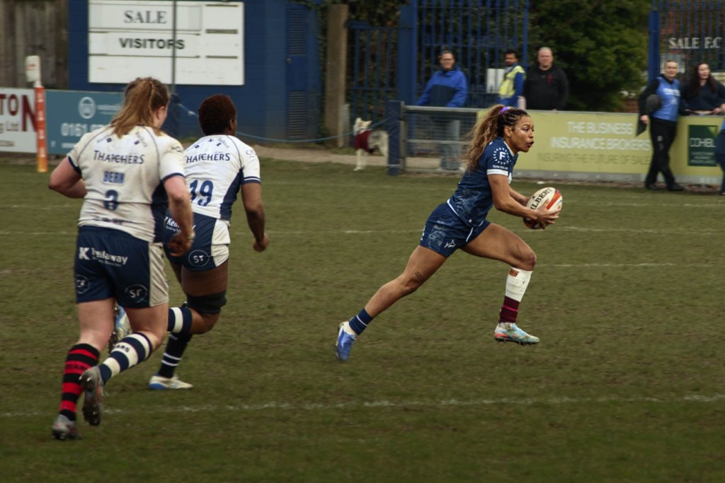 Action shot of a rugby player in blue running with the ball, two opponents in white jerseys chasing from behind on a grassy field.