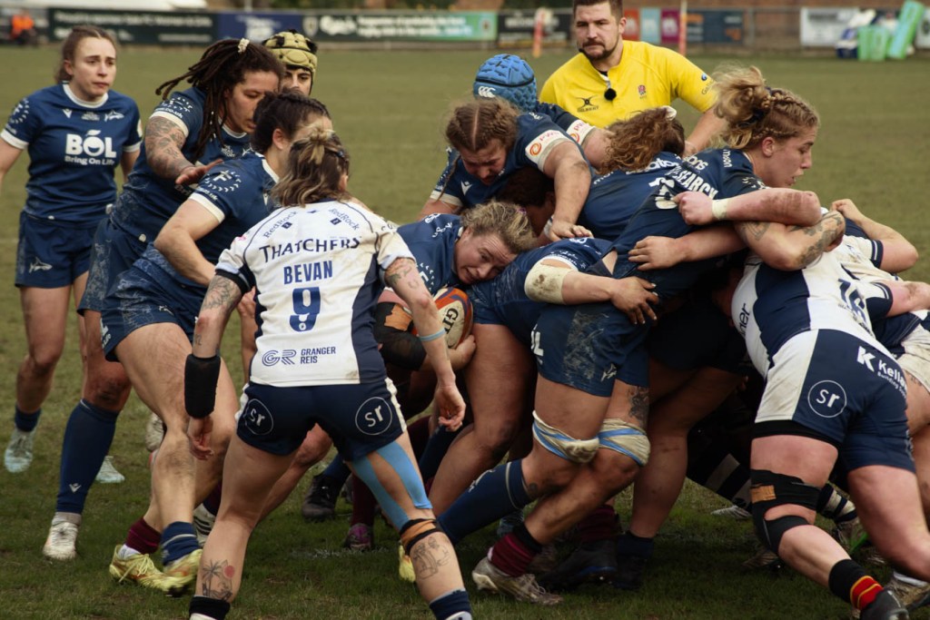 A women's rugby match in action, showing players from both teams engaged in a maul. The scene captures the intensity and physicality of the game on a grassy field.