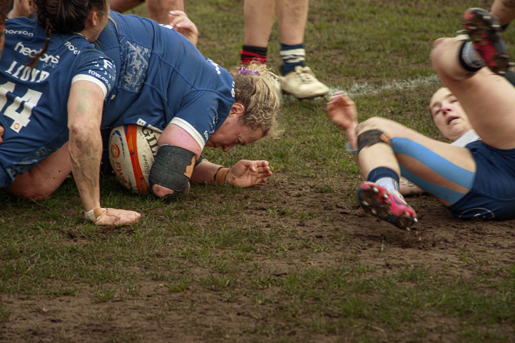 A close-up of players in blue rugby uniforms engaging in a tackle on a muddy field, with one player extending out to score a try.